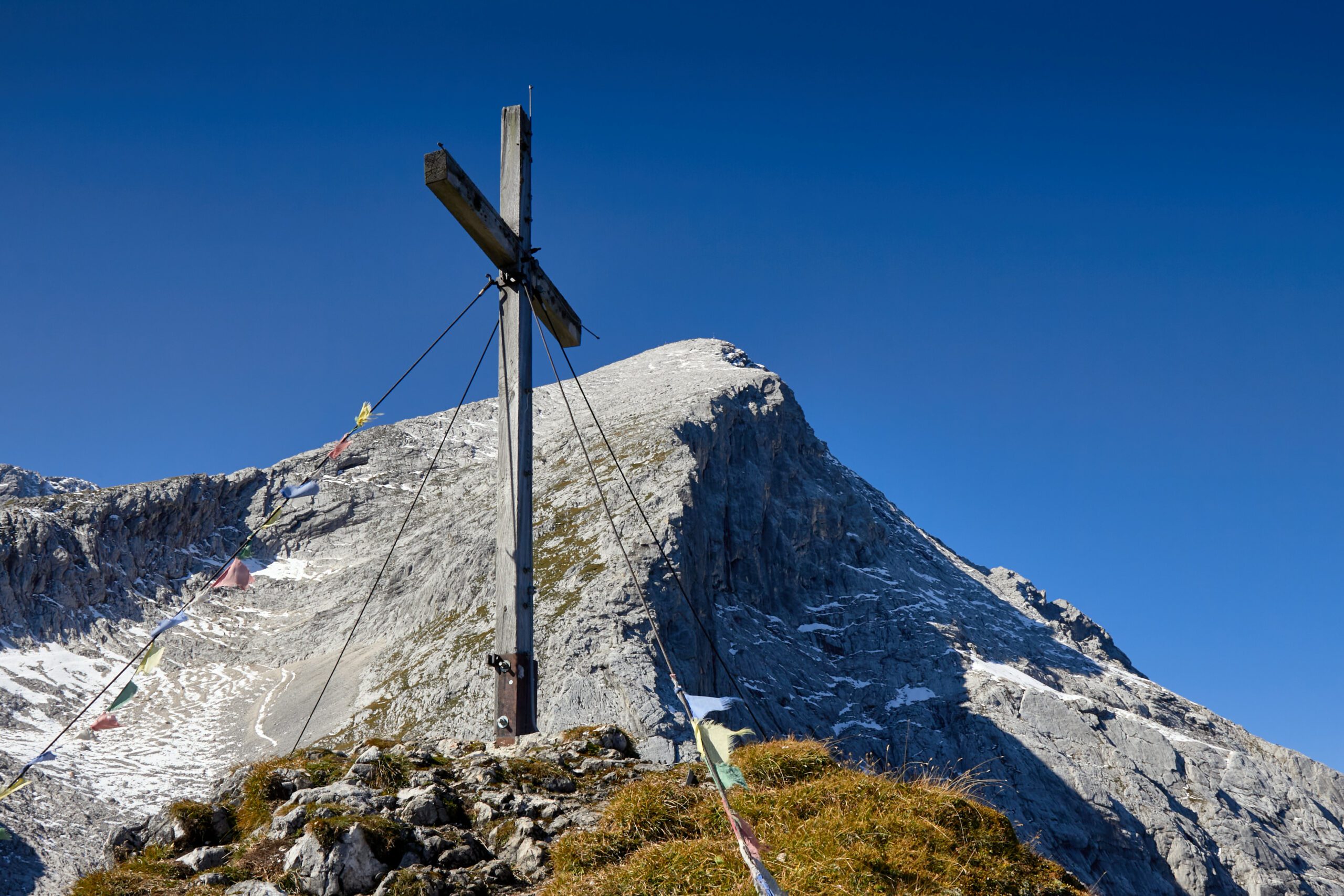 Im Vordergrund steht das Gipfelkreuz des Bernadeinkopfs. Im Hintergrund ragt imposant die Alpspitze vor strahlend blauem Himmel empor.