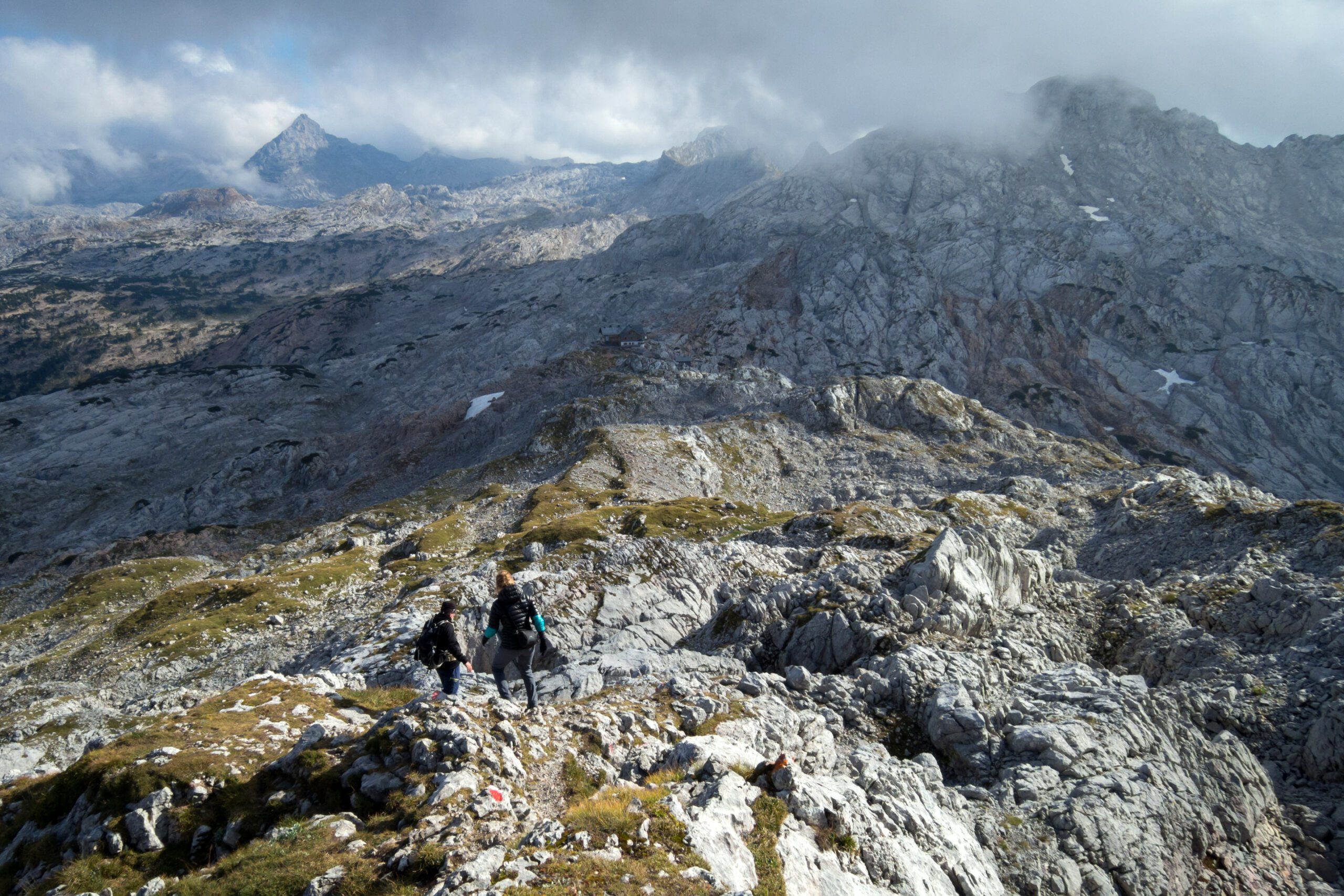 Blick über das Steinerne Meer, Felsen ziehen sich durch die Ebene, zwei Wanderer gehen zu einer Hütte, die noch weit weg ist.